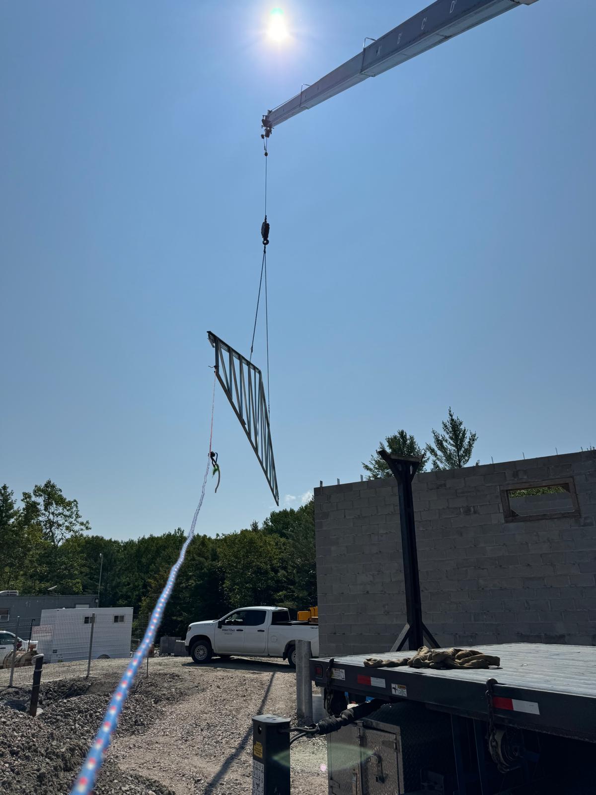 Crane hoisting a steel roof truss over a block wall